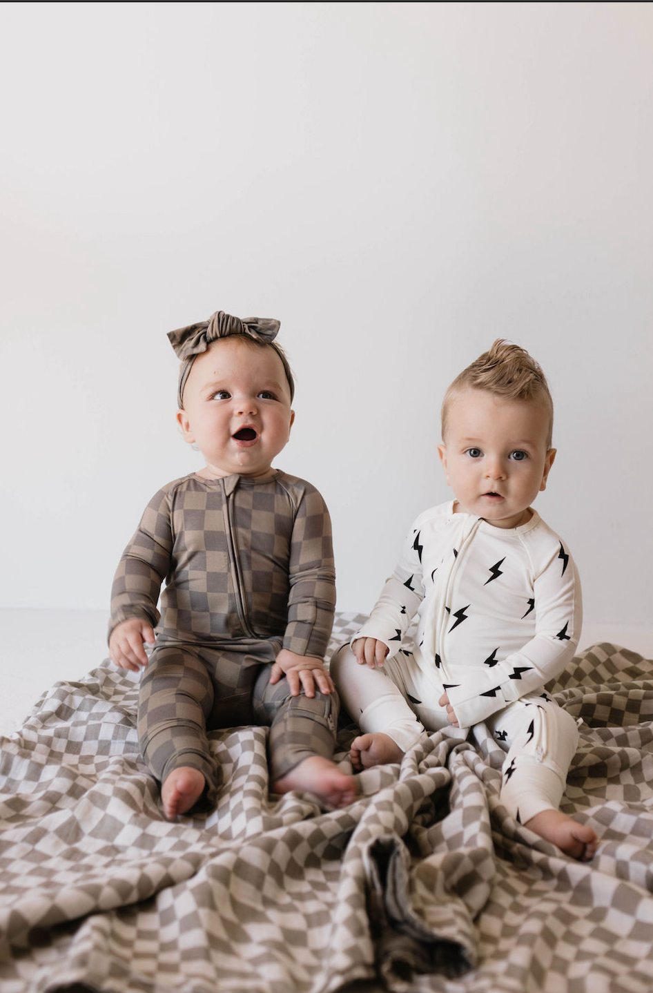 Two babies sit on a checkered blanket against a plain white background. The baby on the left, dressed in a brown checkered onesie and an adjustable Bamboo Head Wrap from forever french baby, looks cozy. The baby on the right sports a white onesie with black lightning bolt prints as both gaze towards the camera.