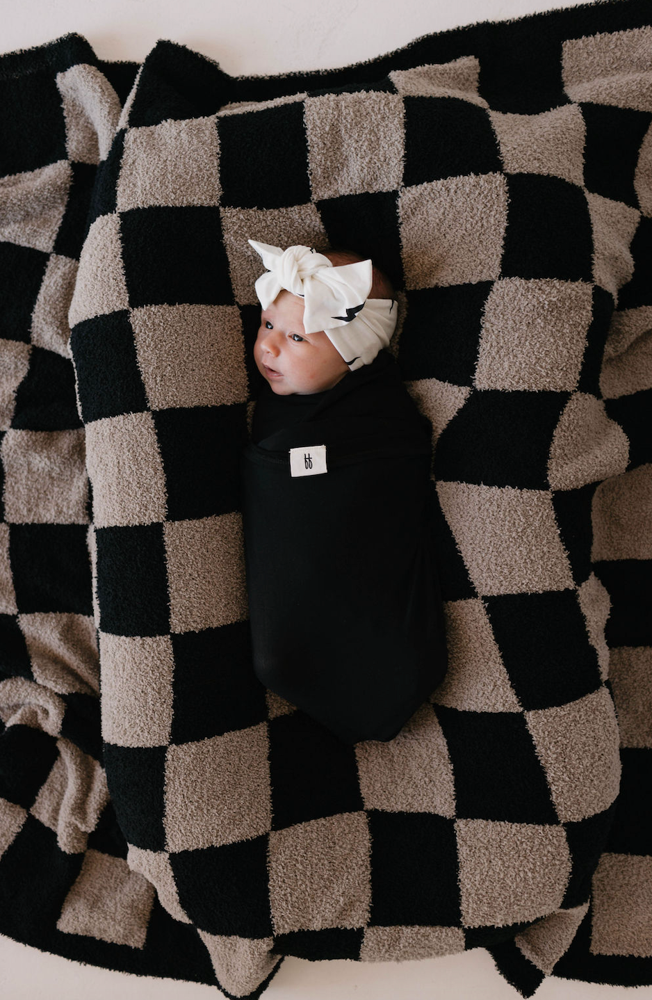 A baby wrapped in a black blanket is lying on a black and beige checkerboard-patterned blanket, adorned with the Bamboo Head Wrap | White + Black Bolt by forever french baby. The setting radiates coziness, evoking a sense of warmth and comfort.