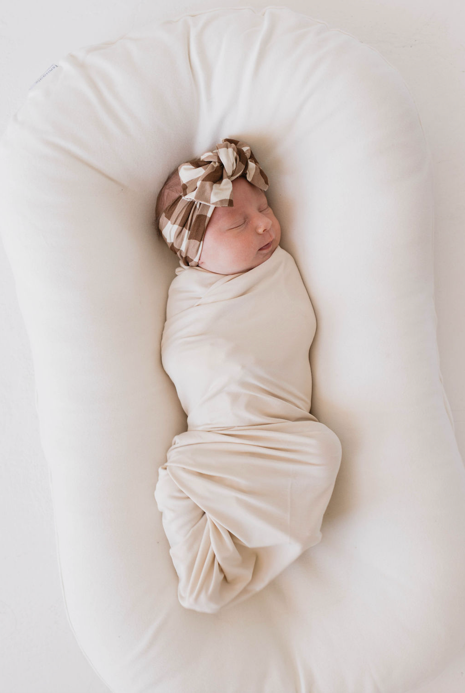 A newborn baby is wrapped tightly in a soft cream-colored swaddle, lying on an oval-shaped cushion. The baby wears a beige and white patterned Bamboo Head Wrap | Groovy Gingham by forever french baby with a bow and appears to be peacefully sleeping. The background is white, creating a clean, serene scene.