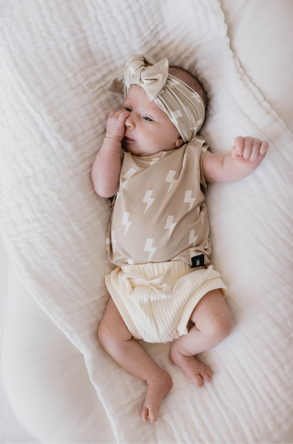 A newborn baby lies on a white blanket, wearing a matching beige headband and romper with a lightning bolt pattern, along with cream-colored shorts. The baby has a relaxed expression and one hand near their mouth, perfectly accessorized with the Bamboo Head Wrap in Tan & Cream Bolt by forever french baby.