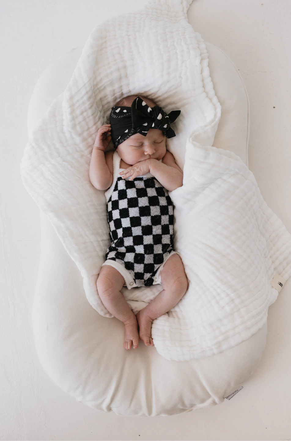 A baby sleeps on a white cushion with a soft, white blanket. The little one is wearing a black and white checkered outfit and the Bamboo Head Wrap in Charcoal & White ff Smile from forever french baby, featuring an adjustable fit with a charming bow. The baby lies on their back with one arm bent and resting near their face.