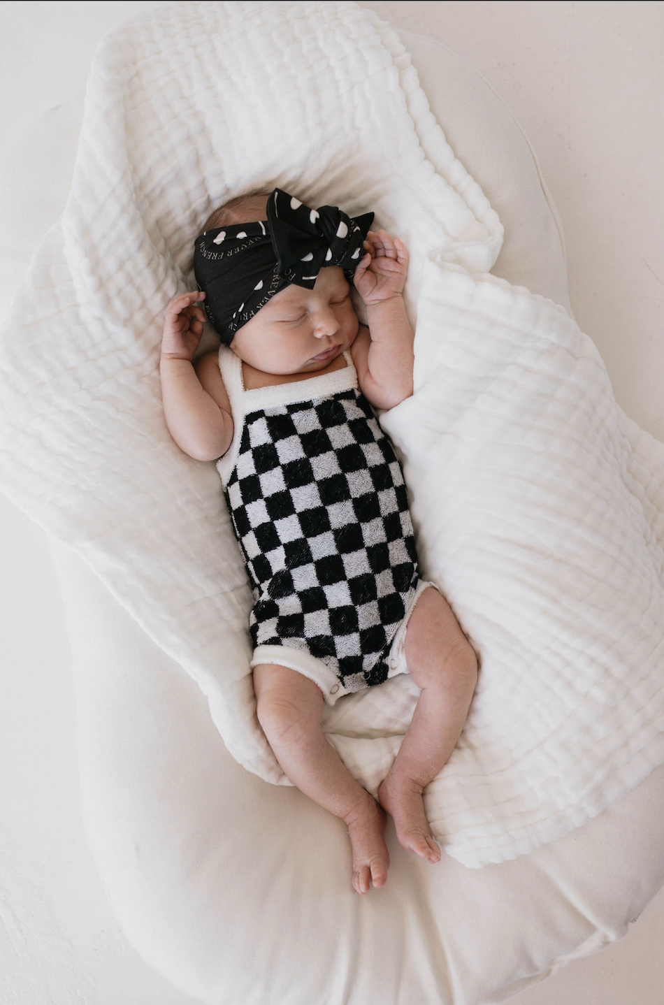 A baby sleeps on a white, textured blanket, wearing a black and white checkered onesie and an adjustable Bamboo Head Wrap in Charcoal & White from forever french baby, featuring a bow. The baby's arms are bent upward, hands resting near its head, with eyes closed, giving a peaceful appearance.