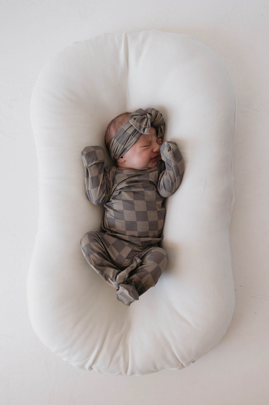 A newborn baby sleeps on a cushioned white pillow. Dressed in a matching checkered onesie and an adjustable Bamboo Head Wrap in Faded Brown Checker by forever french baby, the baby’s hands are near its face, peacefully resting. The soft, gentle bamboo material ensures comfort for babies and toddlers alike.