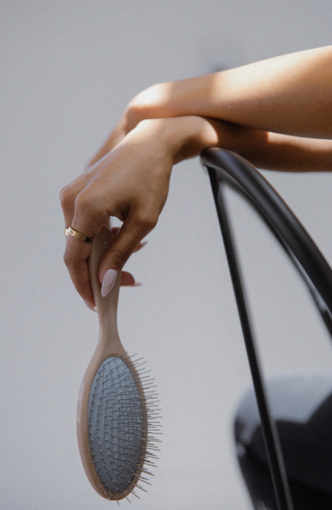 A person's hand, adorned with light-colored nail polish and a gold ring, rests on the back of a chair while holding the Glow Beauty Hair Brush from forever french baby. The brush features slender metal bristles and a light-colored handle. The background is softly lit and out of focus.