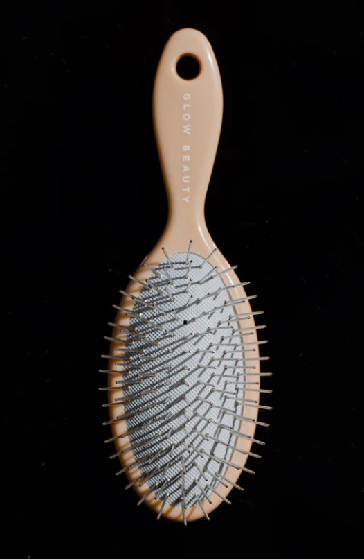 A beige, oval-shaped hairbrush with metal bristles and a plastic handle, branded as "forever french baby: Glow Beauty Hair Brush." The black background makes this detangling brush the focal point of the image.