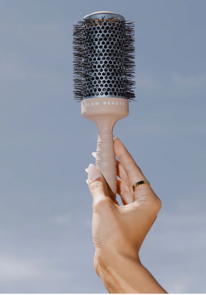 A hand with long manicured nails holds up a Glow Beauty Hair Brush by the handle against a clear blue sky. The brush features a mesh-like circular barrel with black bristles and "forever french baby" written on the handle.