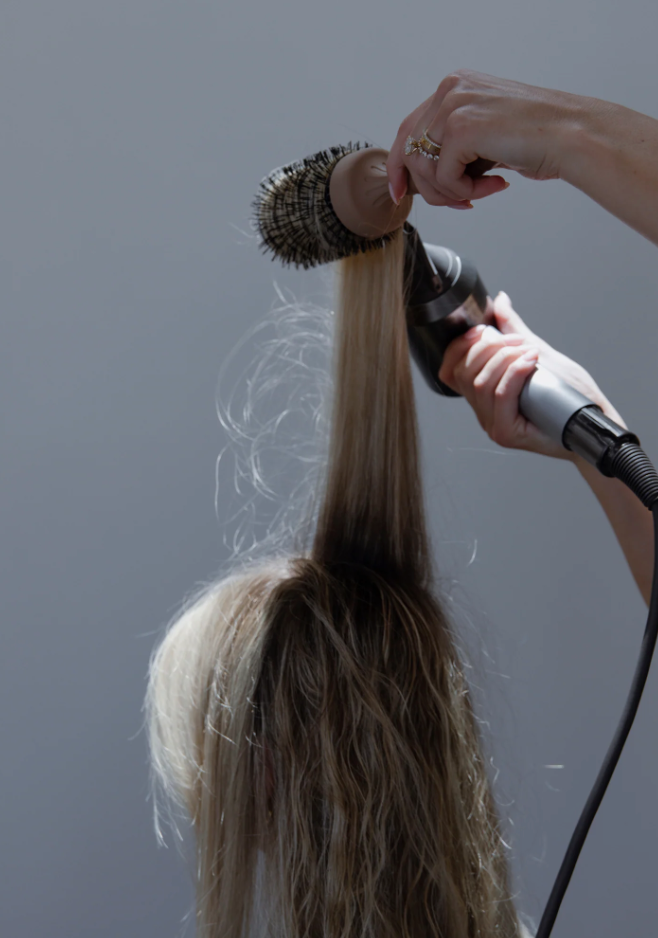 A person using the Glow Beauty Hair Brush by forever french baby and a hairdryer to style another person's long, straight blonde hair. The hairstylist lifts a section of hair with the Glow Beauty Hair Brush while aiming the hairdryer at it.