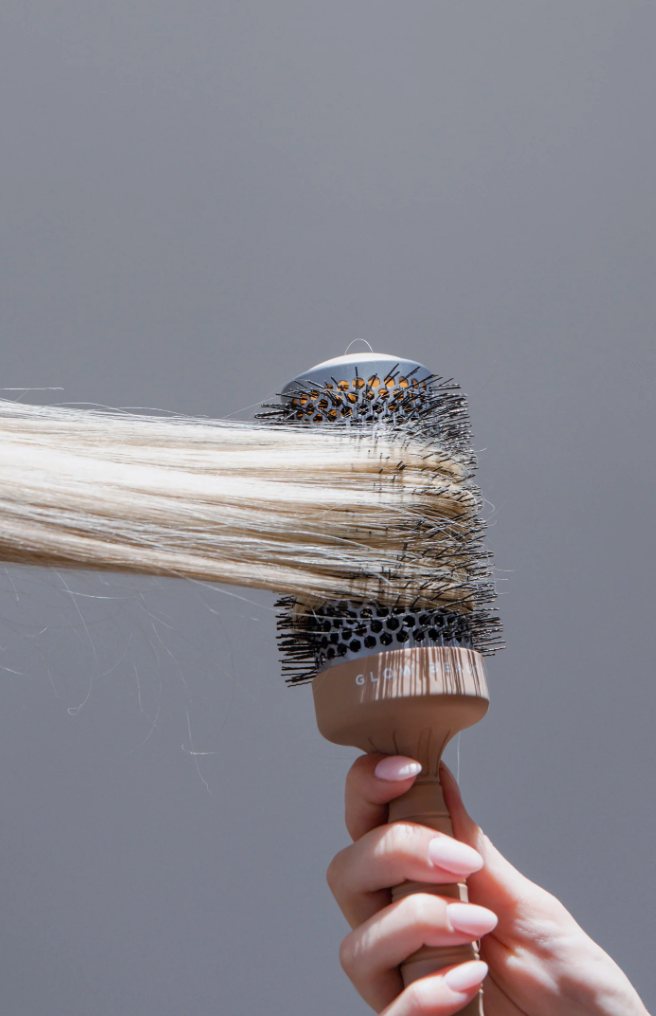 A person holding a Glow Beauty Hair Brush by forever french baby, brushing long, straight, platinum blonde hair against a gray background. The brush features a wooden handle, a metal barrel, and black bristles with orange tips.