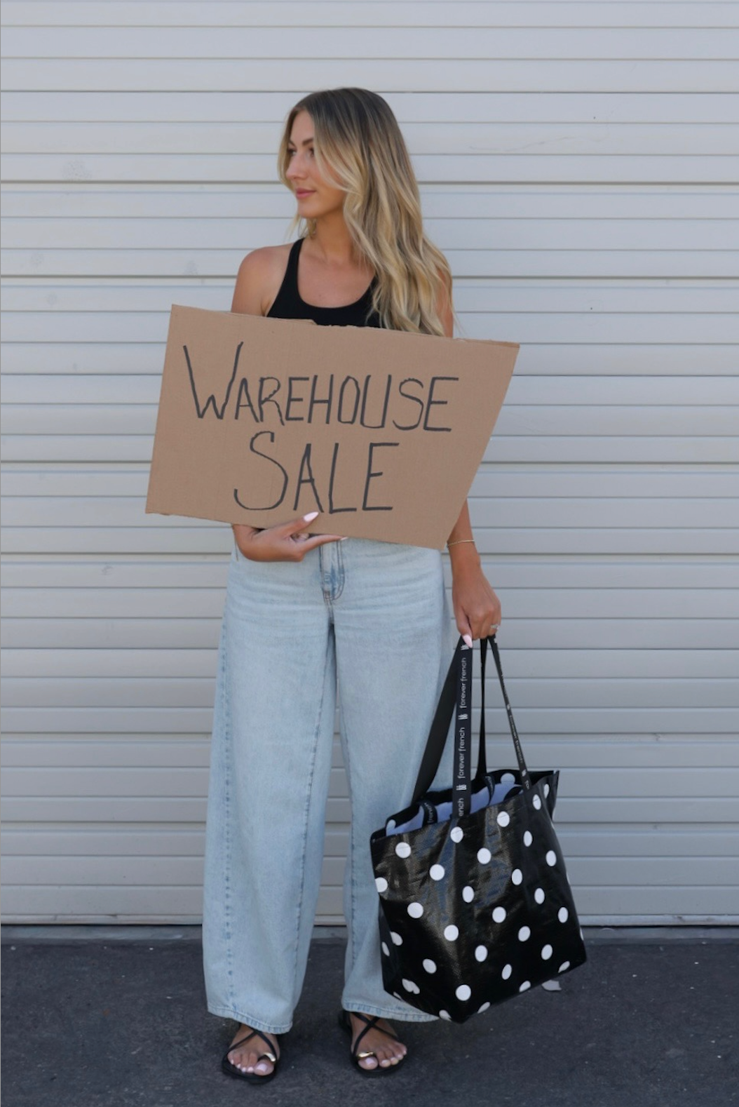 A woman in a black tank top and wide-leg jeans stands in front of a white metal door, holding a cardboard “Warehouse Sale” sign and the ff Warehouse Sale Tote Bag | B&W Gingham by forever french baby.