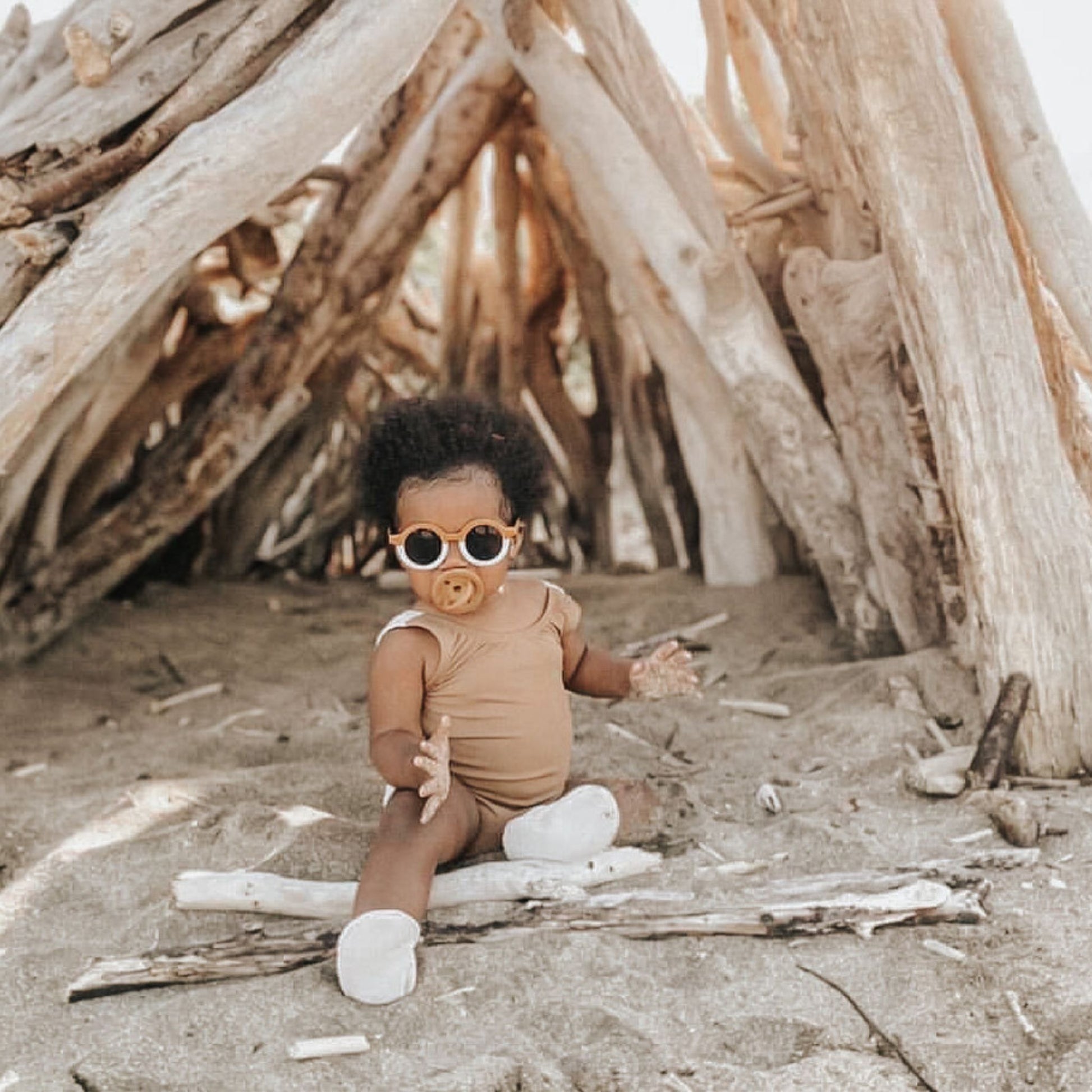 A baby, sporting round sunglasses and a beige onesie, is seated on sandy ground before a structure fashioned from sizable driftwood pieces. With a Modern Pacifier in Caramel by forever french baby in their mouth and white socks on their feet, the baby relishes the sunny beach atmosphere.