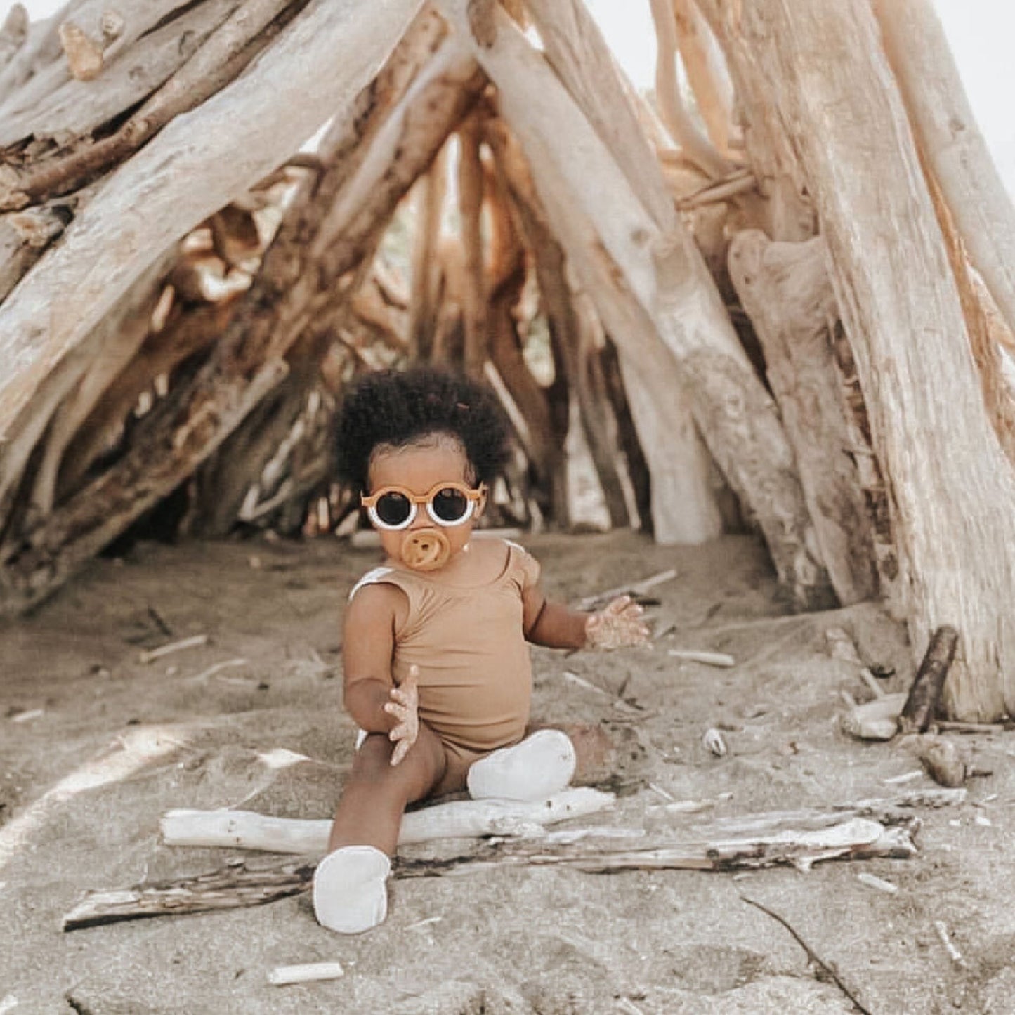 A baby wearing sunglasses, a **Modern Pacifier in Moss by forever french baby**, and a brown onesie sits on the sand in front of a structure made from driftwood. The baby has curly hair and white shoes. The driftwood structure arches above, creating a rustic, playful environment perfect for showcasing essential silicone designs.