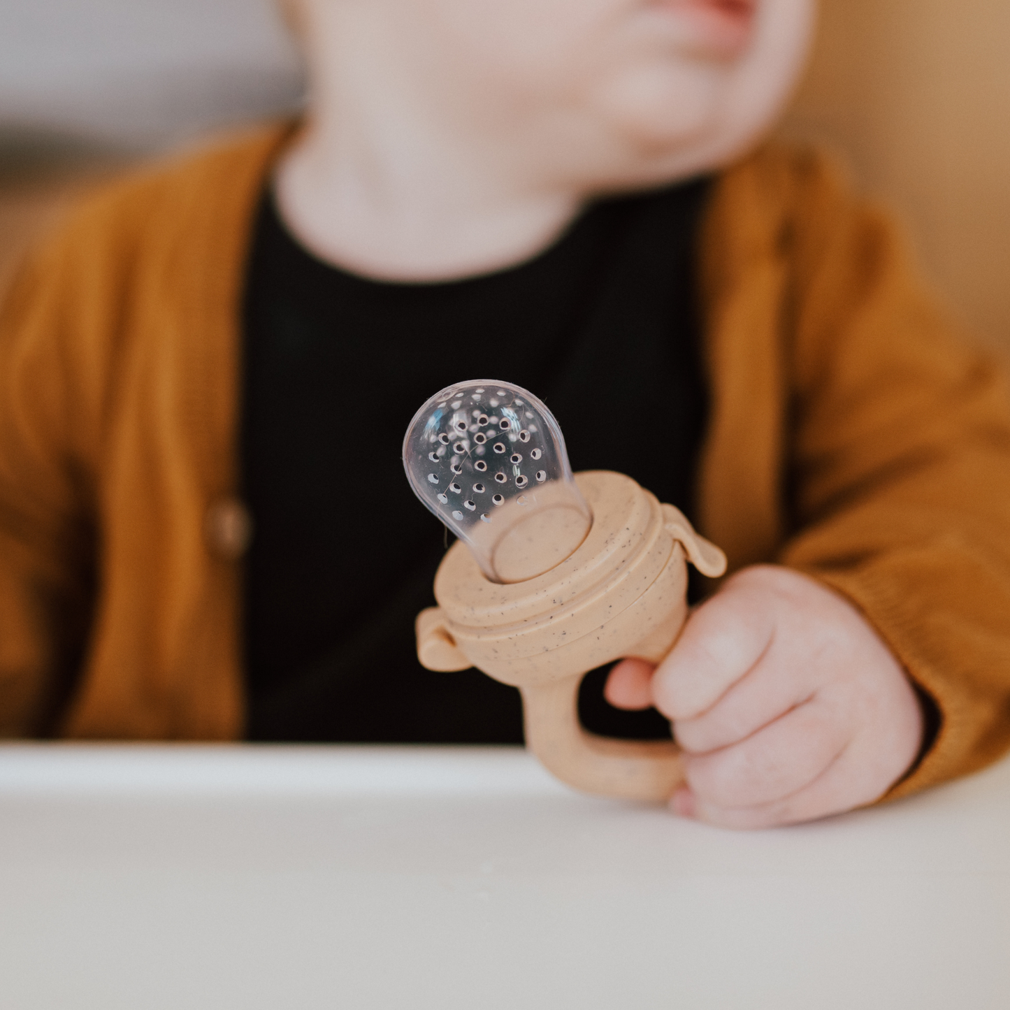 A baby wearing a brown cardigan and black shirt is holding the forever french baby Modern Mod Feeder, which features a clear beaded top, while seated at a white surface. The background is blurred, drawing attention to the baby's hand and the toy.