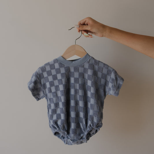 A hand holds a hanger with the Terry T-Shirt Romper | Tradewinds by forever french baby—this short-sleeved, blue and gray checkered romper is ideal for matching family outfits, displayed against a plain light-colored wall.