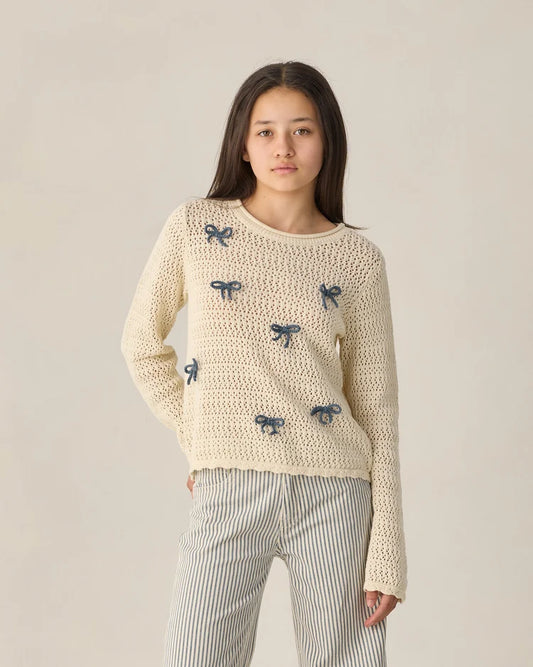 A young person wears the Rylee + Cru Teen Crochet Knit Pullover | Bows—featuring a cream crochet design with blue bow details—and striped pants, standing against a plain light background while looking at the camera with a neutral expression.