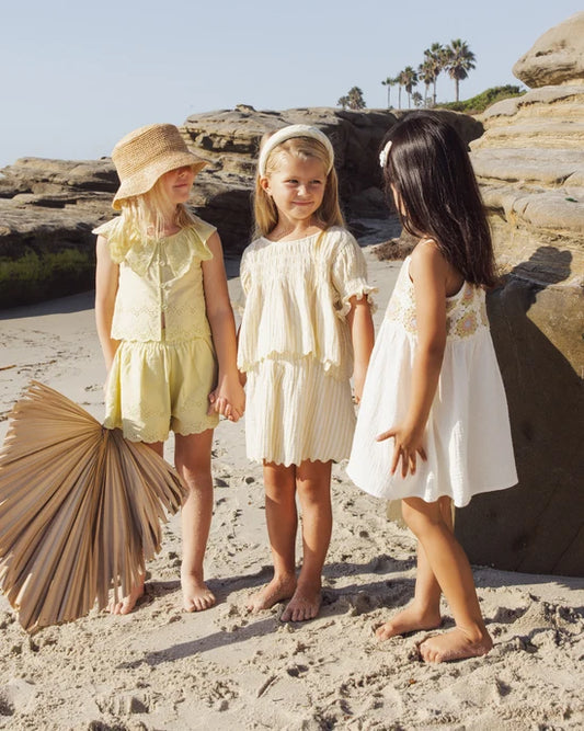 Three young girls stand barefoot on a sandy beach near rocks, talking and smiling. One wears the Colette Top in Citron Stripe by Rylee + Cru, its light summer fabric featuring delicate embroidered scallop edges. One of the girls holds a large palm fan, while palm trees and a blue sky stretch across the background.