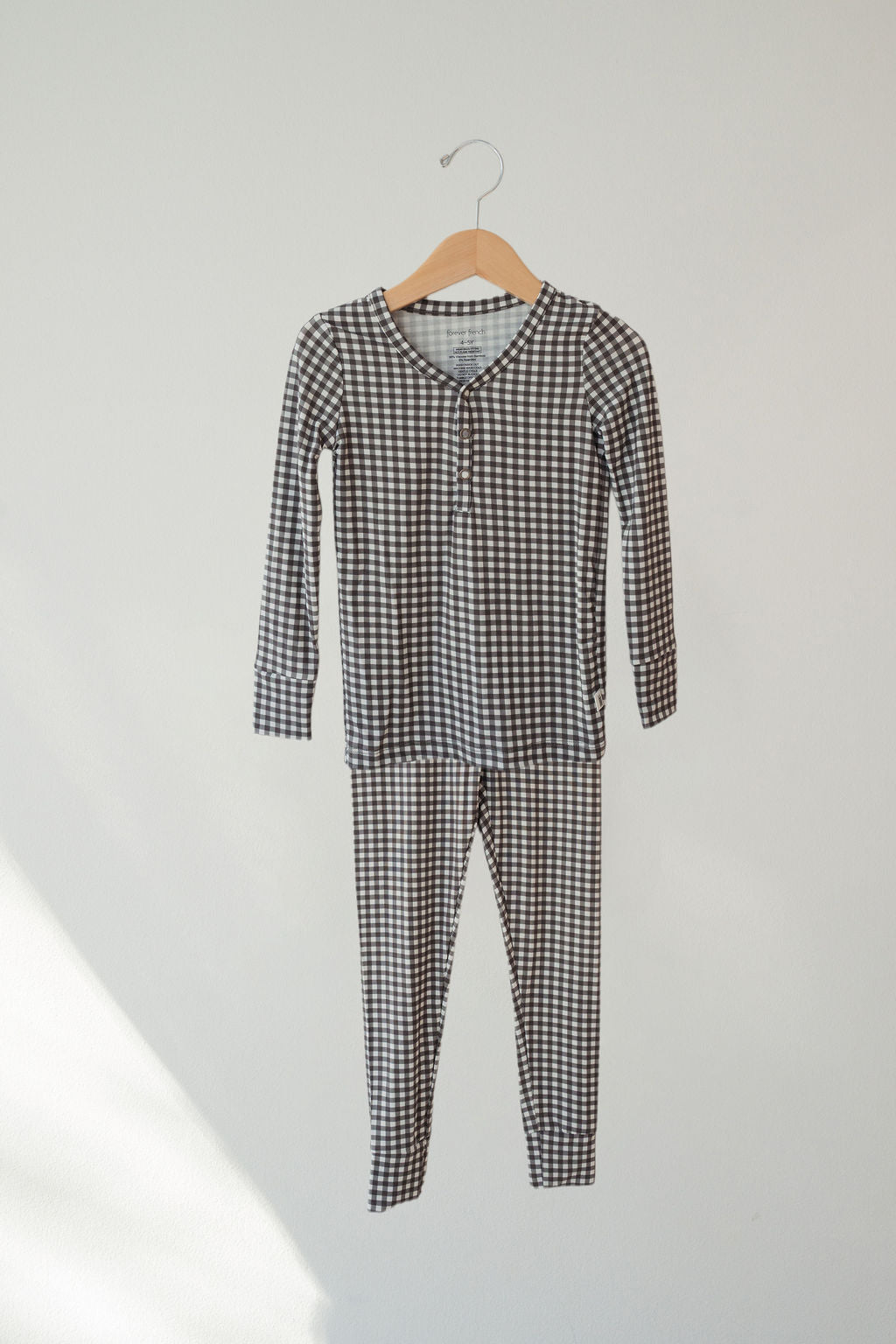 The forever french baby Bamboo Two Piece Pajamas in Green Gingham, made from breathable bamboo, hangs on a wooden hanger against a plain, light-colored wall.