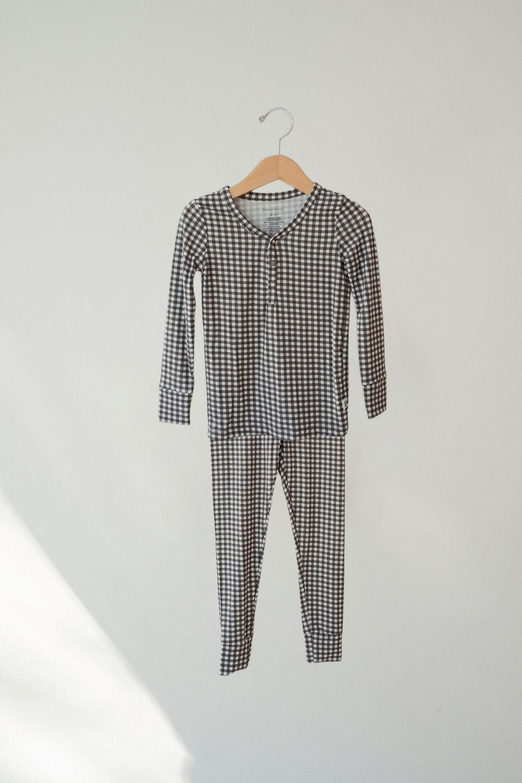 The forever french baby Bamboo Two Piece Pajamas in green gingham, a hypoallergenic kids set, hangs on a wooden hanger against a plain light-colored wall.