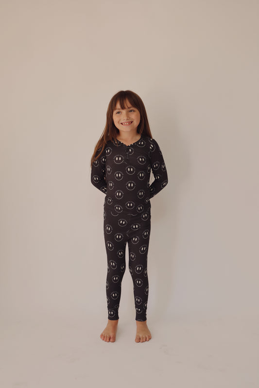 A young child with long brown hair stands barefoot, smiling and wearing the Bamboo Two Piece Pajamas in Charcoal & White ff Smile by forever french baby. The child stands against a plain off-white background.