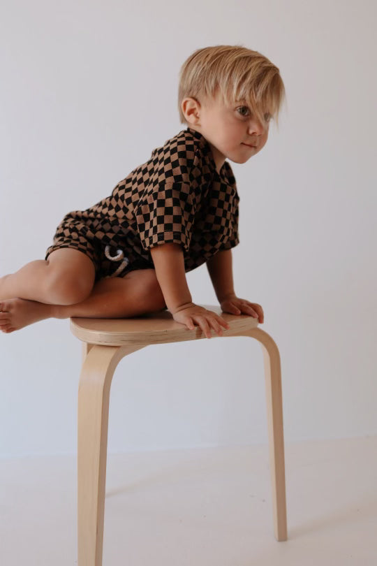 A blonde toddler, dressed in a Ribbed Short Set | Black & Brown Checkerboard by forever french baby, is sitting on a light wooden stool, leaning slightly forward with one leg bent. The minimalistic setting features a plain white background.