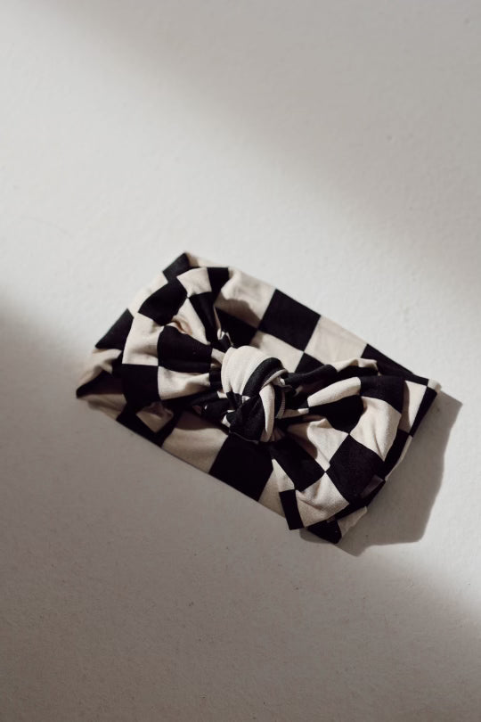 A folded piece of fabric with a black and white checkerboard pattern is fashioned into a bow on a light, textured surface. The lighting casts subtle shadows, accentuating the fabric’s folds and knot—a perfect Bamboo Head Wrap | Black Checkerboard from forever french baby for babies and toddlers.