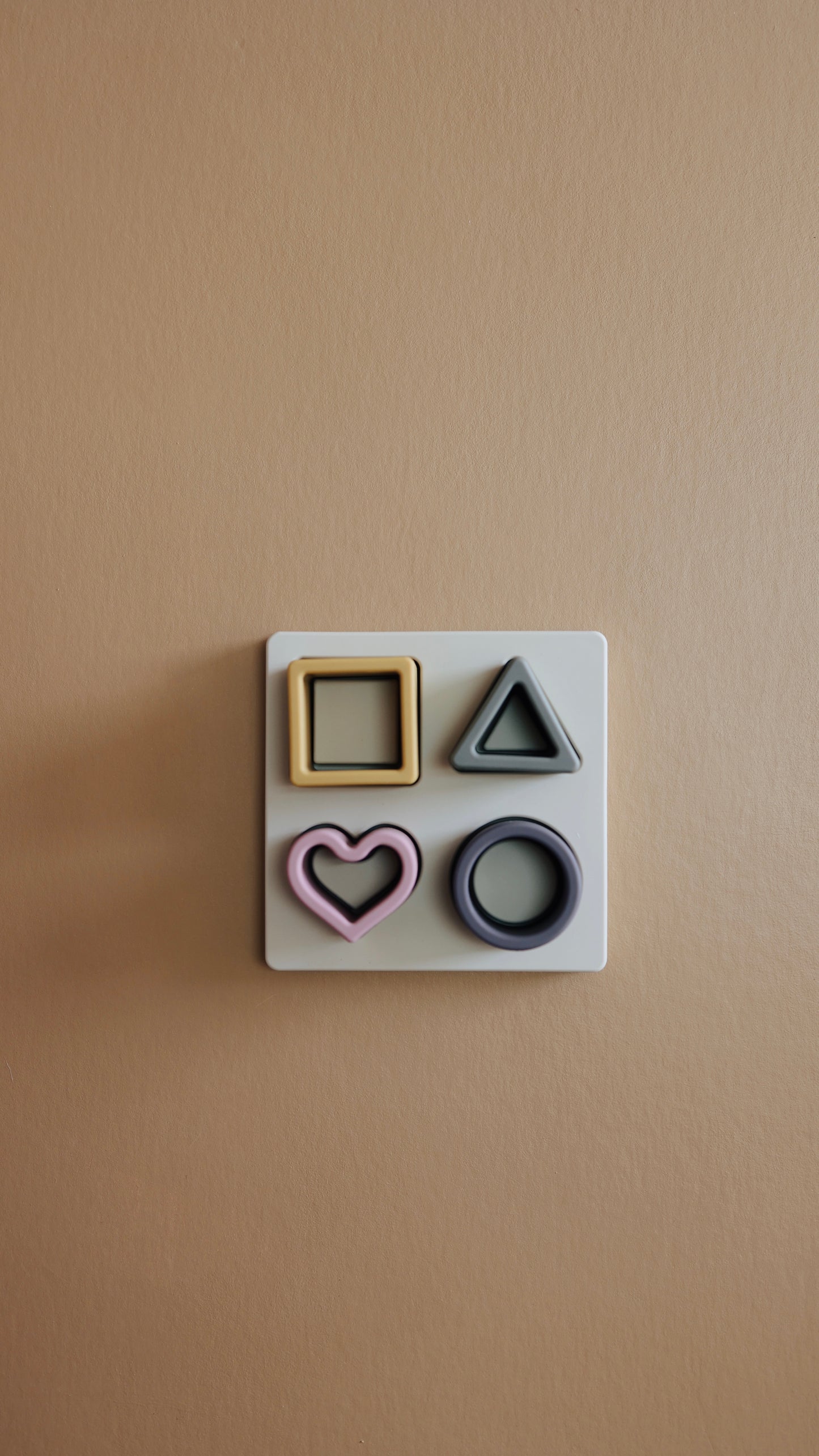 A beige wall features the forever french baby Silicone Puzzle | Toy with four geometric shapes placed: a yellow square, a pink heart, a green triangle, and a blue circle. Made from non-toxic BPA-free silicone, these shapes are neatly organized, with each fitting into a corresponding recess on the board.