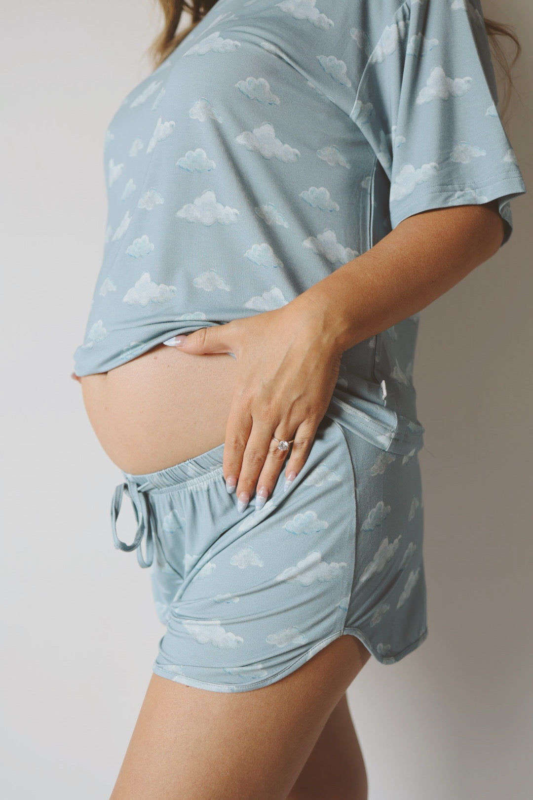 A pregnant woman stands sideways, gently touching her bare baby bump while wearing Kendy 3's Women's Bamboo Short Set | Duke—light blue, breathable pajamas with a cloud pattern.