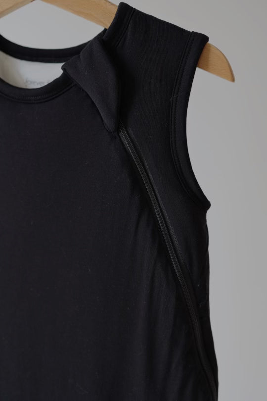 A close-up of the Bamboo Sleep Sack in Midnight Black by forever french baby, displayed on a wooden hanger, highlights its zippered detail along the side. The fabric, reminiscent of breathable bamboo material, appears soft and stretchy, making it suitable for both casual and active wear. The background is plain and neutral.