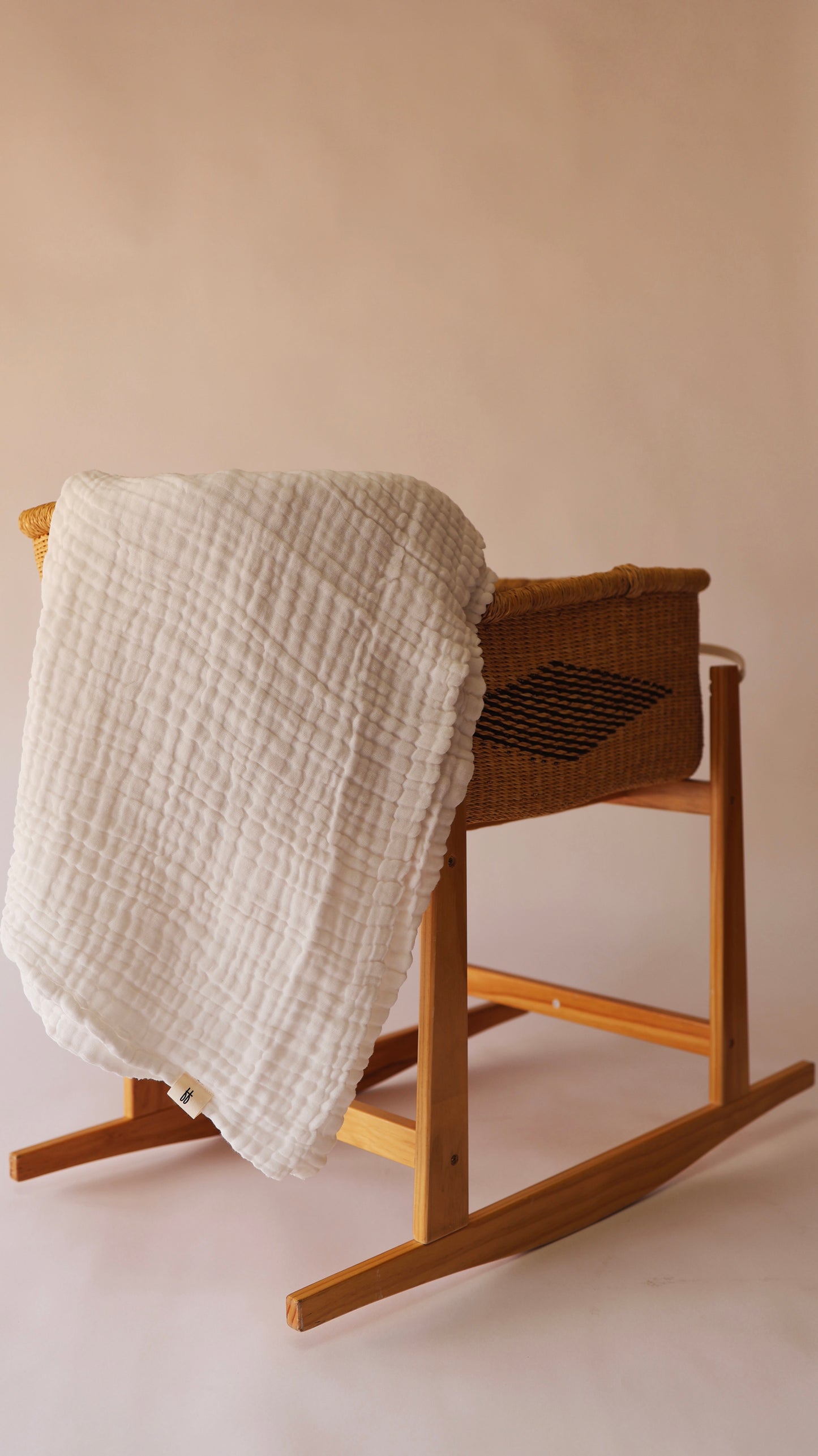 A wicker rocking bassinet from forever french baby, adorned with the Crib Sheet | Cloud that features a clean, white quilt draped over its side, is set against a plain beige background. The bassinet's simple wooden frame beautifully complements the quilt's textured grid-like pattern. For lasting quality, remember to wash the crib sheet with like colors and tumble dry on low.