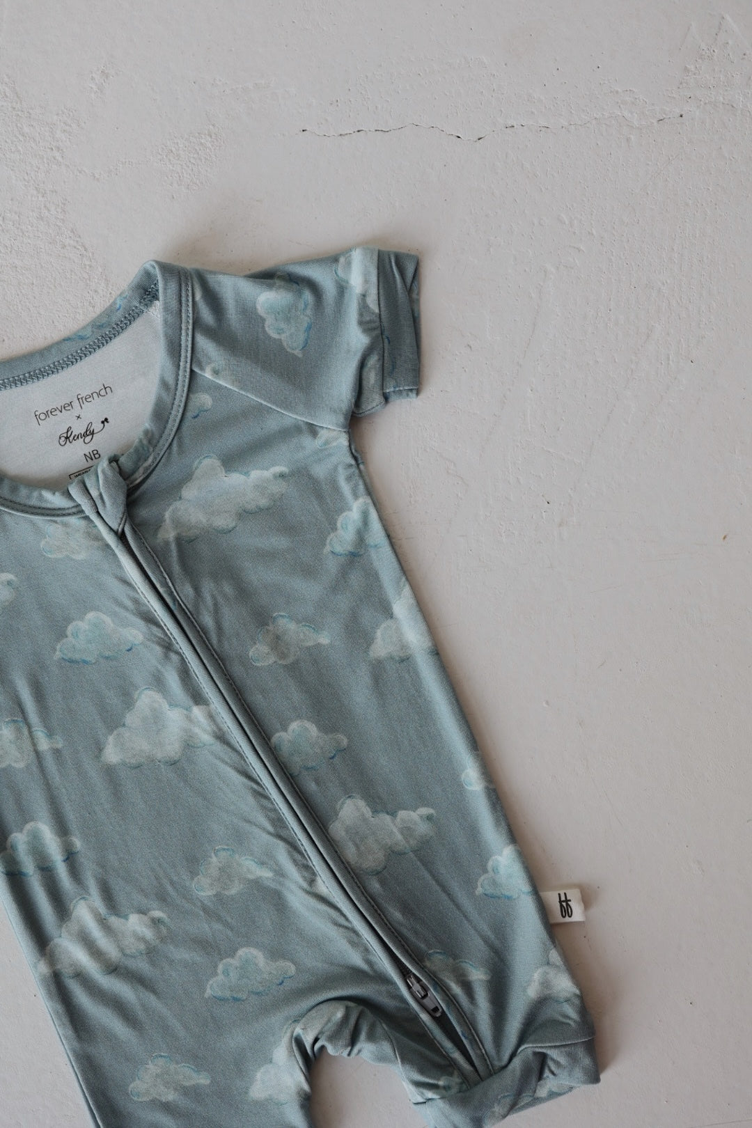 The Kendy 3 Bamboo Zip Shortie | Duke is a light blue, short-sleeved baby romper with a white cloud pattern and front zipper, made from breathable fabric for ultimate comfort and shown laid flat on a light textured surface.