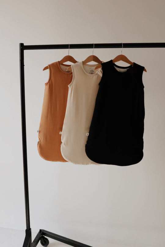 Three sleeveless Bamboo Sleep Sacks from forever french baby, in various shades (brown, cream, black), hang on wooden hangers from a black metal clothing rack. The backdrop is a minimalistic, light-colored wall. These Bamboo Sleep Sacks | Desert Sunset are eco-friendly wearable blankets that offer both style and sustainability for your baby's comfort.