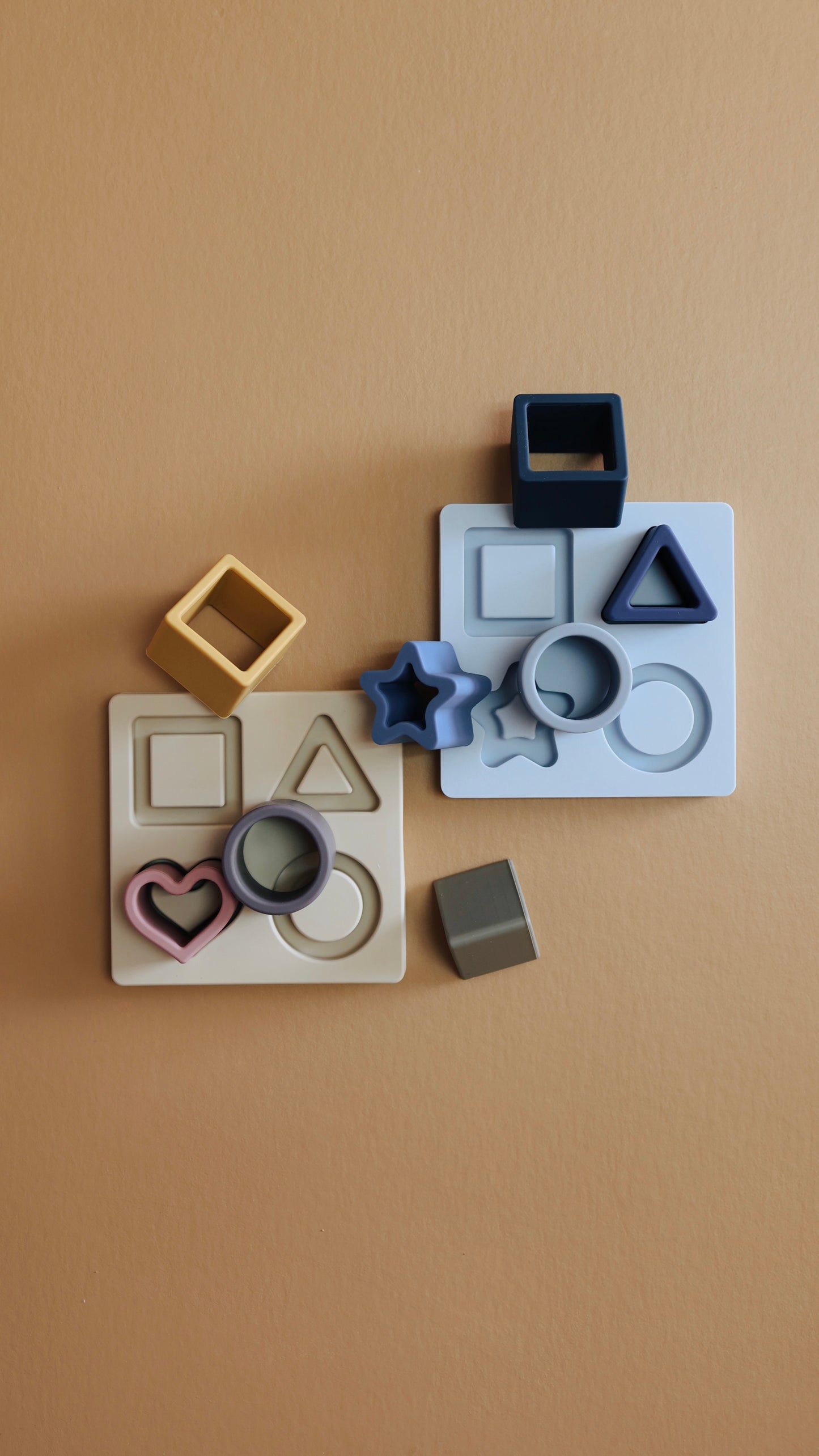 Two sets of forever french baby Silicone Puzzles are arranged on a smooth beige surface. These assorted pastel-colored shape sorters feature stars, triangles, squares, circles, and hearts. Made from non-toxic BPA-free silicone, these safe and durable toys make bath time fun and educational. The shapes fit into corresponding color-coordinated holes to enhance learning.