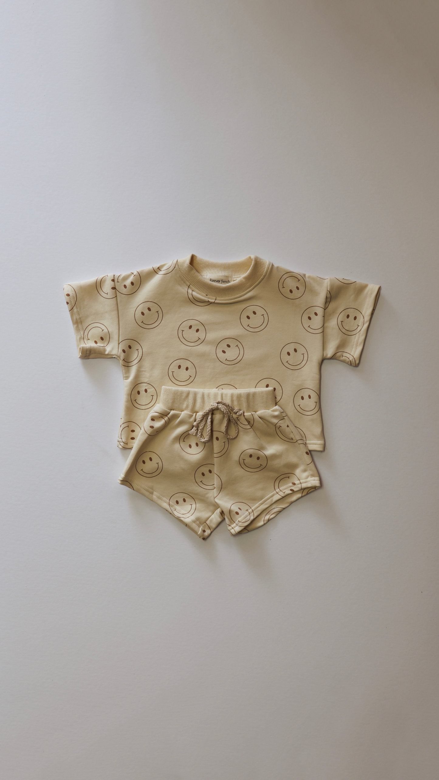 The Just Smile Ivory short set from forever french baby includes a beige baby outfit featuring a matching short-sleeve shirt and shorts adorned with smiling faces. The shirt has a round neck, and the shorts come with an elastic waistband and drawstring. Perfect as a transitioning piece for any season, this delightful ensemble is laid flat on a light surface.