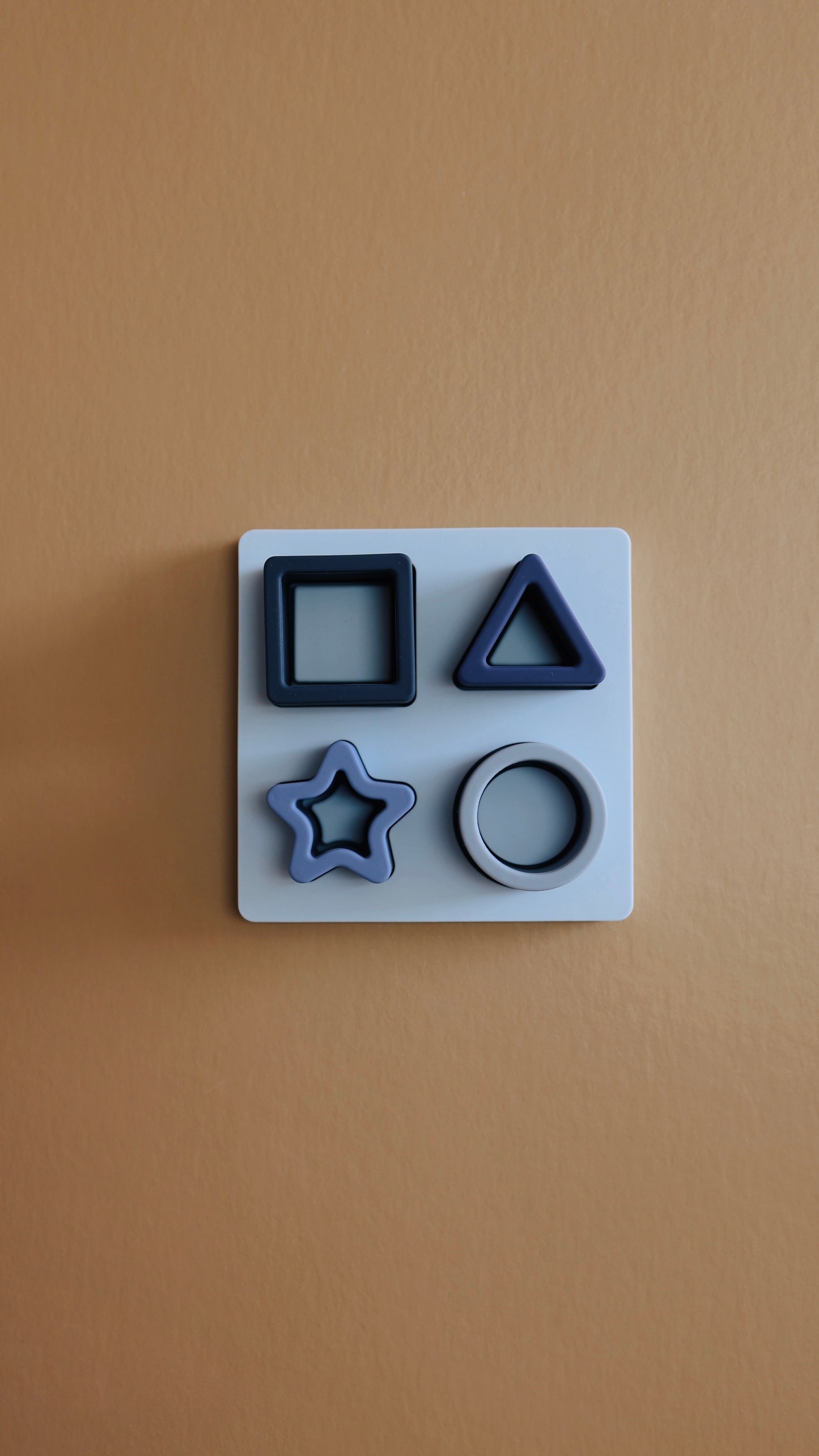 A white square-mounted board from the forever french baby Silicone Puzzle | Toy is attached to a beige wall, showcasing four shape indentations: a square, triangle, star, and circle, each filled with light blue molds made of non-toxic BPA-free silicone.