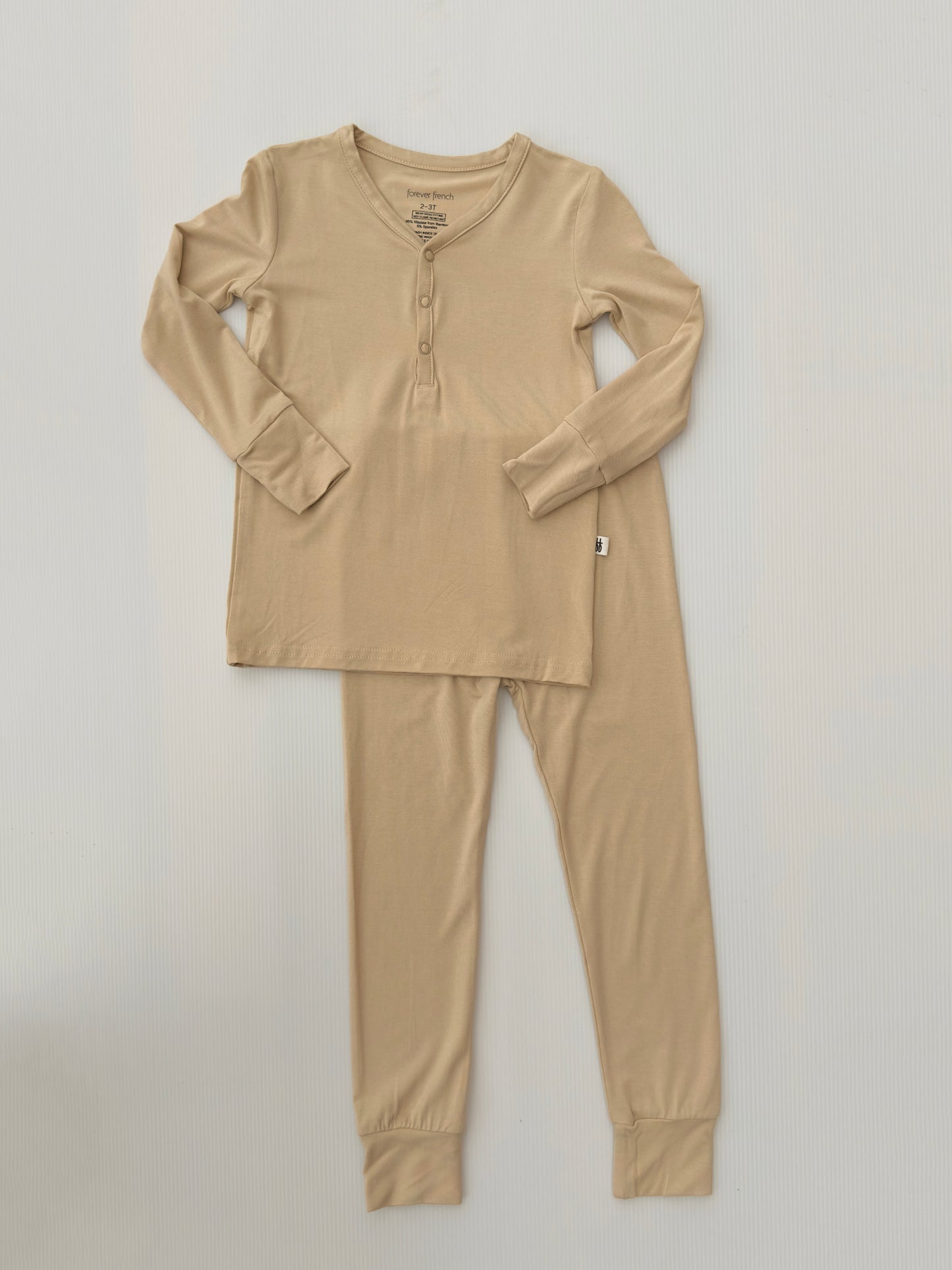 The "Bamboo Two Piece Pajamas | Limestone" from forever french baby, a beige-colored pajama set made from breathable bamboo fabric, featuring a long-sleeved, button-up top and matching pants, is laid flat against a white background. The top has three buttons on the neckline. The pants have an elastic waistband and cuffs at the ankles.