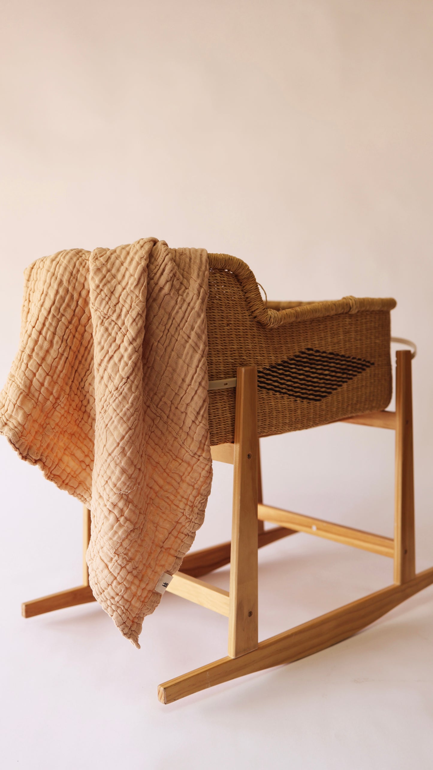 A quilted newborn blanket in a warm latte color drapes over the side of a wicker baby bassinet with wooden rocking legs. This bassinet, from forever french baby, stands empty against a plain, light background, creating a minimalist and cozy atmosphere.