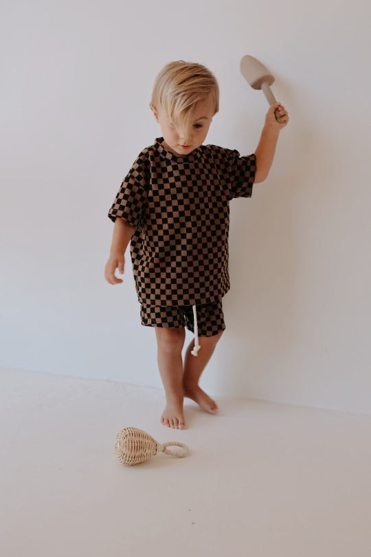 A young child, likely in the 0-9 years age range, with blonde hair is standing barefoot, dressed in a Ribbed Short Set | Black & Brown Checkerboard by forever french baby. The child is holding a wooden rattle and looking down at another wicker rattle lying on the floor.