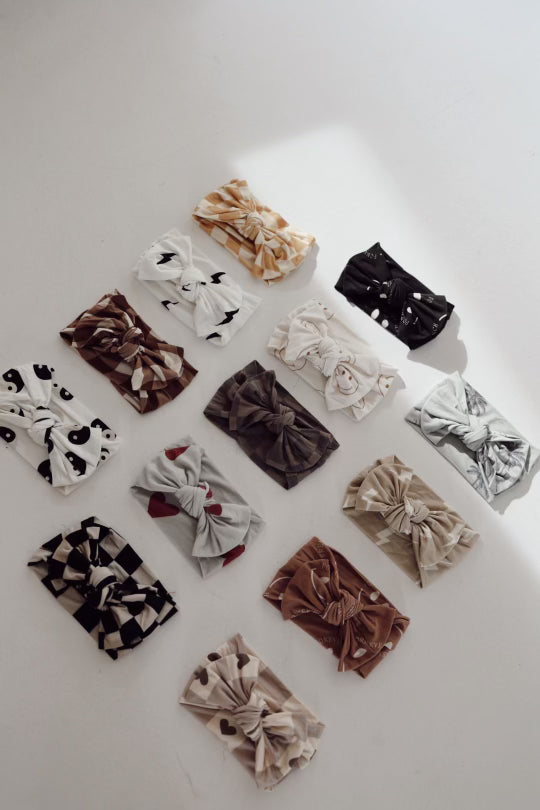 A collection of 14 adjustable Bamboo Head Wraps by forever french baby, arranged on a white surface, featuring various patterns such as polka dots, abstract shapes, and animal prints in shades of white, black, brown, beige, and gray. Each wrap is tied in the center with a bow.