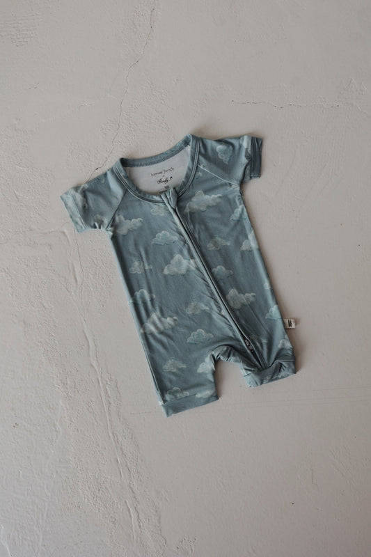 The Kendy 3 Bamboo Zip Shortie | Duke is a short-sleeved, light blue baby romper with a cloud pattern and front zipper, made from breathable fabric and shown laid flat on a textured light surface.