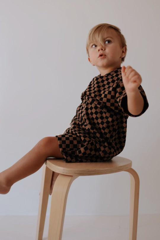 A small child with blond hair sits on a light wooden stool against a plain background. The child is wearing a Ribbed Short Set | Black & Brown Checkerboard outfit by forever french baby and has one arm raised, looking off to the side with an expression of curiosity.