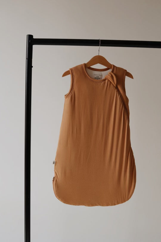 The Bamboo Sleep Sack | Desert Sunset by forever french baby, in a tan and sleeveless design, hangs on a wooden hanger from a black horizontal rod against a plain white background. This eco-friendly sleeping bag features a soft, simple design with a rounded bottom and a small fabric tag on one side.