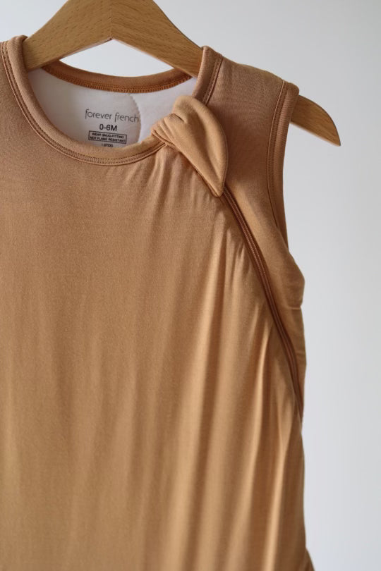 A Bamboo Sleep Sack in a tan hue, designed with a sleeveless cut and featuring a small knotted detail on one shoulder, is hanging on a wooden hanger. This eco-friendly garment from forever french baby is tailored for infants aged 0-6 months, embodying both style and sustainability under the name "Desert Sunset.