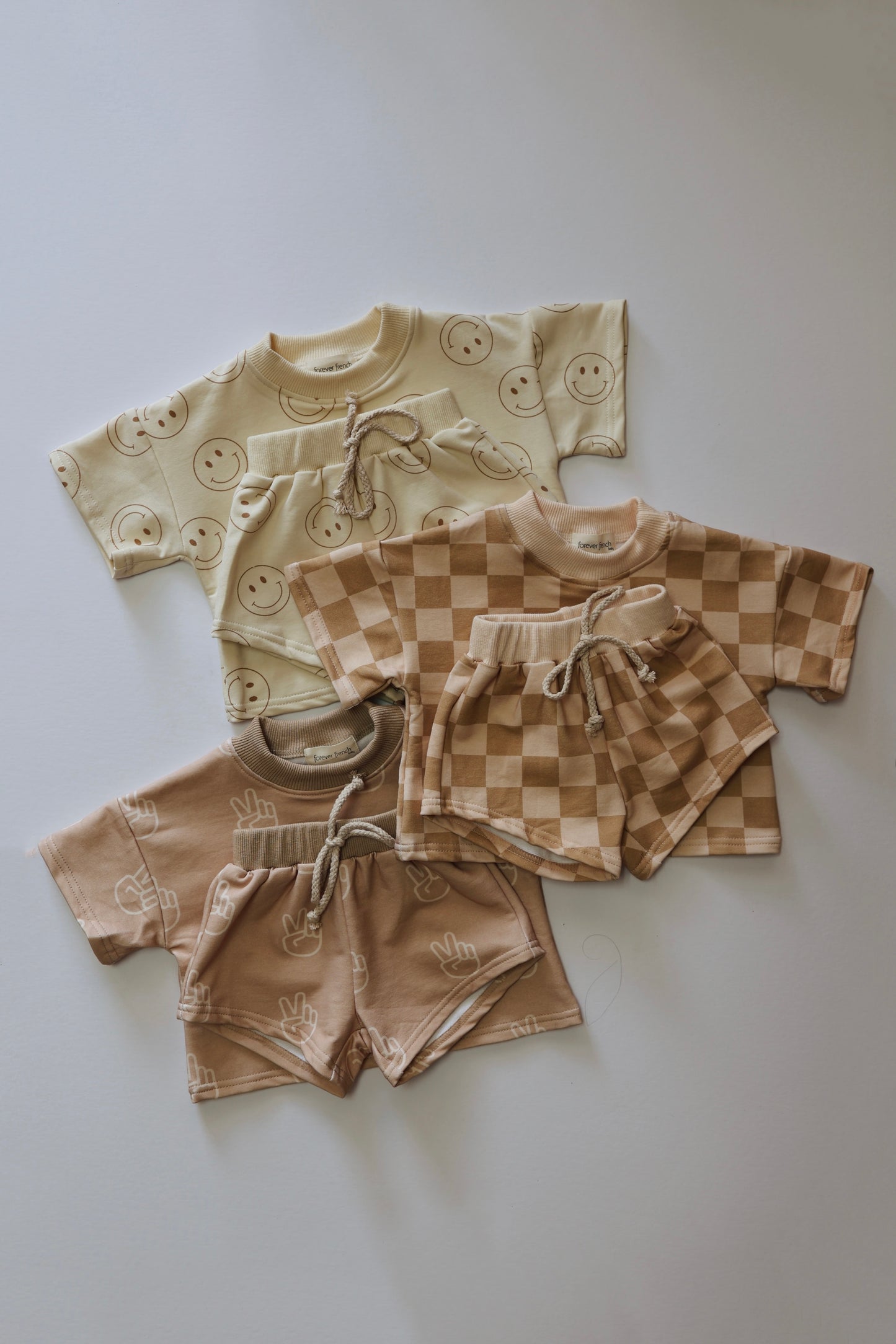 Four sets of the "Short Set | Just Smile Ivory" children's clothing from Forever French Baby are neatly laid out. Each set includes a short-sleeve shirt and matching shorts, featuring delightful designs of smiley faces, checkerboard patterns, and hand symbols in neutral tones of beige and brown—ideal pieces for transitioning between seasons.