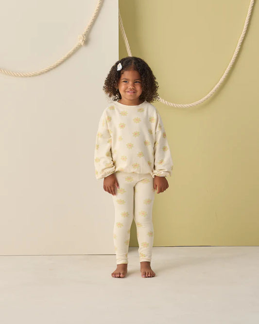 A young child with curly hair, barefoot and smiling, models the Rylee + Cru Spongey Knit Set | Yellow Daisy—a coordinated toddler sweatshirt and leggings adorned with a delightful yellow daisy pattern—against a pale green and cream backdrop decorated with ropes.