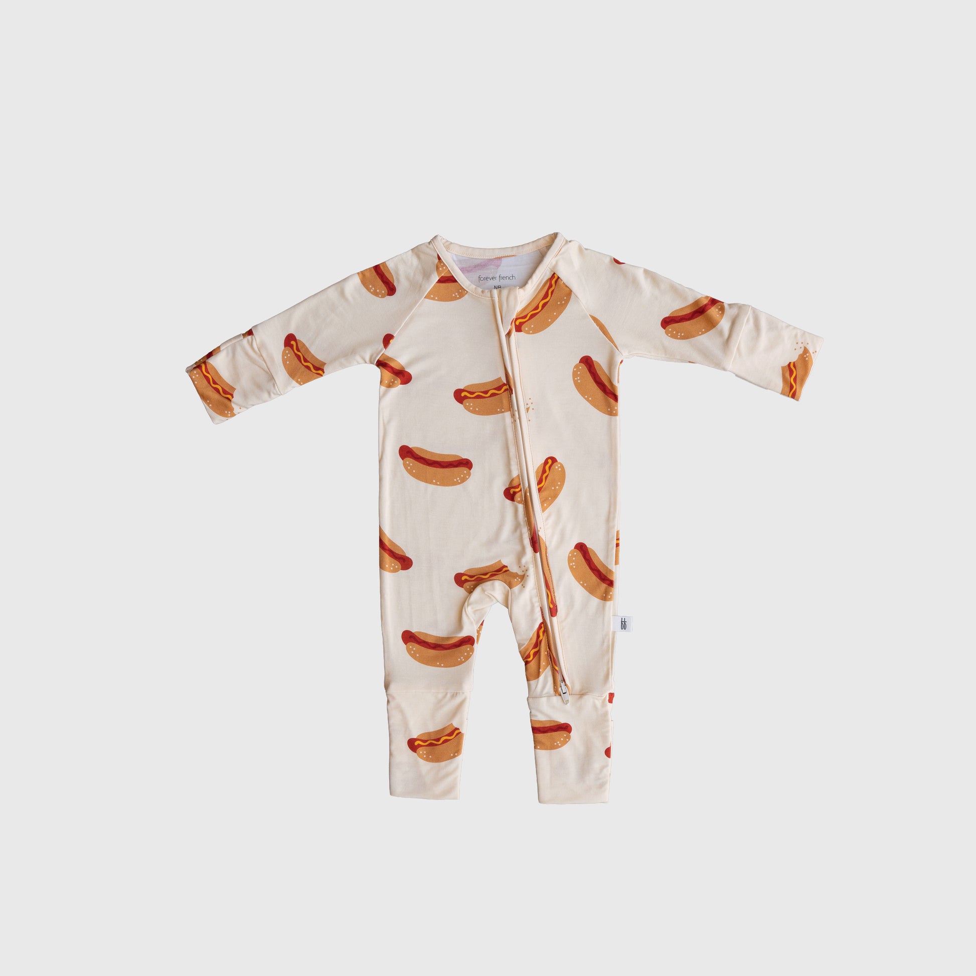 The forever french baby Bamboo Zip Pajamas | Hot Dogs is a cream-colored bamboo viscose baby onesie with a zipper and an all-over hot dog print, shown on a plain light background.