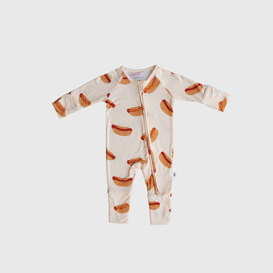 The forever french baby Bamboo Zip Pajamas | Hot Dogs is a cream-colored bamboo viscose baby onesie with a zipper and an all-over hot dog print, shown on a plain light background.