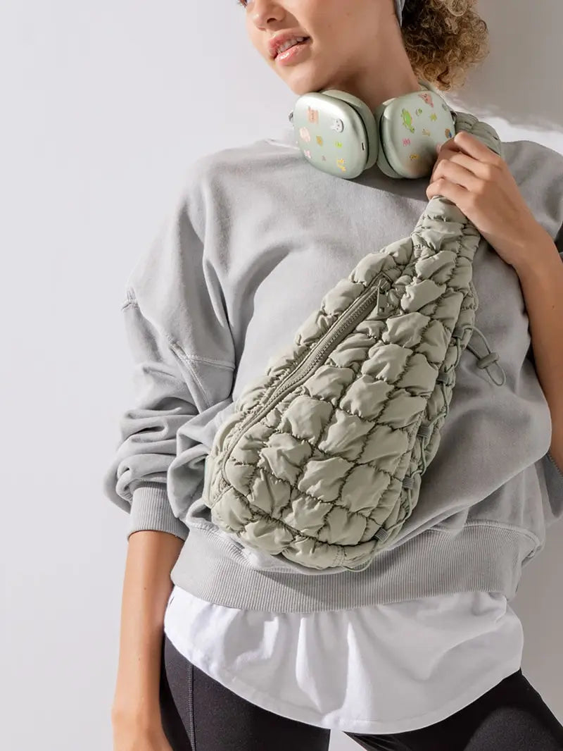 A woman wearing a light grey sweatshirt and white shirt holds the urban daizy Quilted Sling Bag in sage green, perfect for everyday use. She has chunky headphones with a playful, colorful design around her neck. Standing casually, her hand rests on the strap of this stylish accessory, which features a zipper pocket.