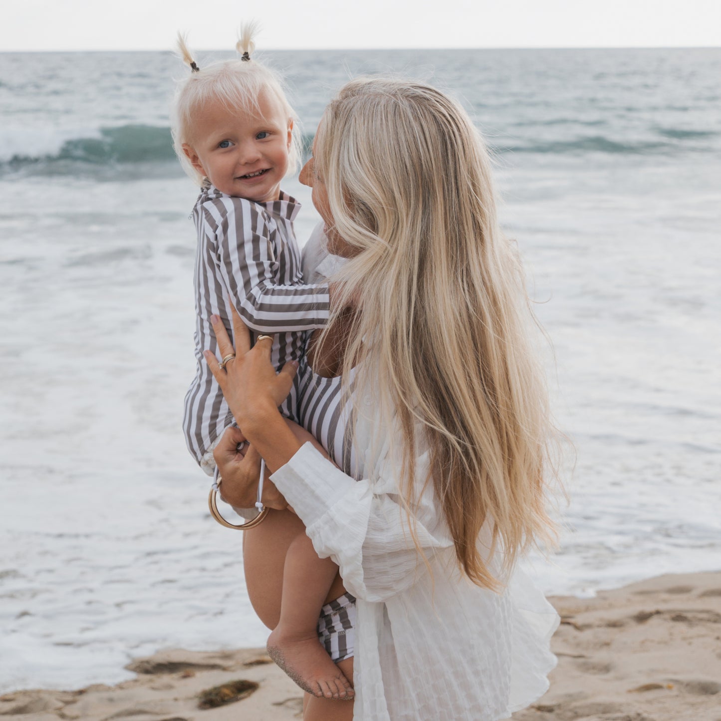 A woman with long blonde hair holds a smiling toddler in blonde pigtails, both on a sandy beach. The toddler wears the Girl's Long Sleeve Swimsuit in Olive Stripe by forever french baby, with waves visible in the background.