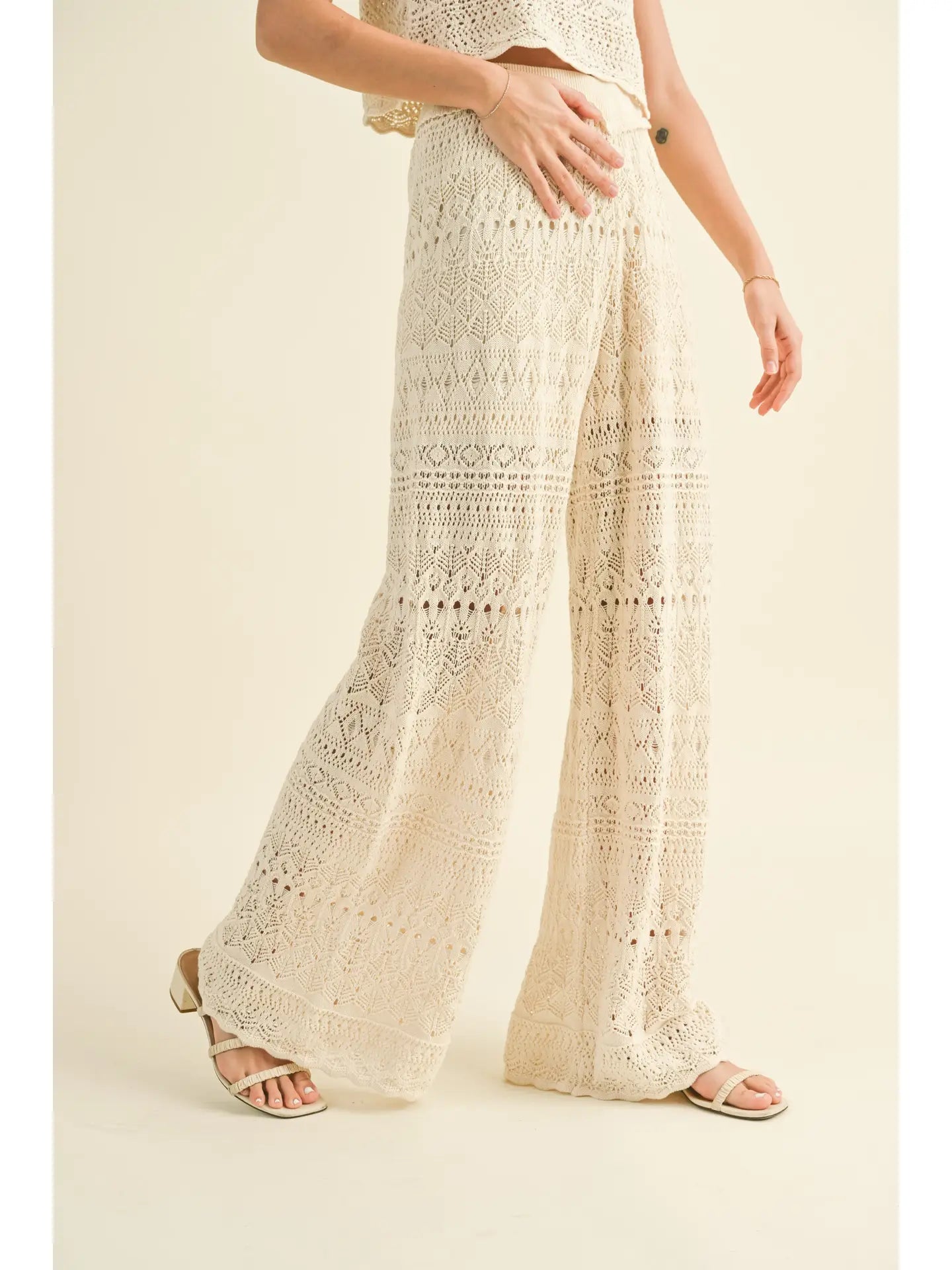 A person is dressed in MIOU MUSE's Crochet Pattern Pants, featuring a high-waisted design in a soft cream cotton with intricate patterns. The ensemble includes a coordinating top and is styled with sandals against a beige backdrop, with the person placing one hand on their waist. The pants necessitate gentle hand wash care.