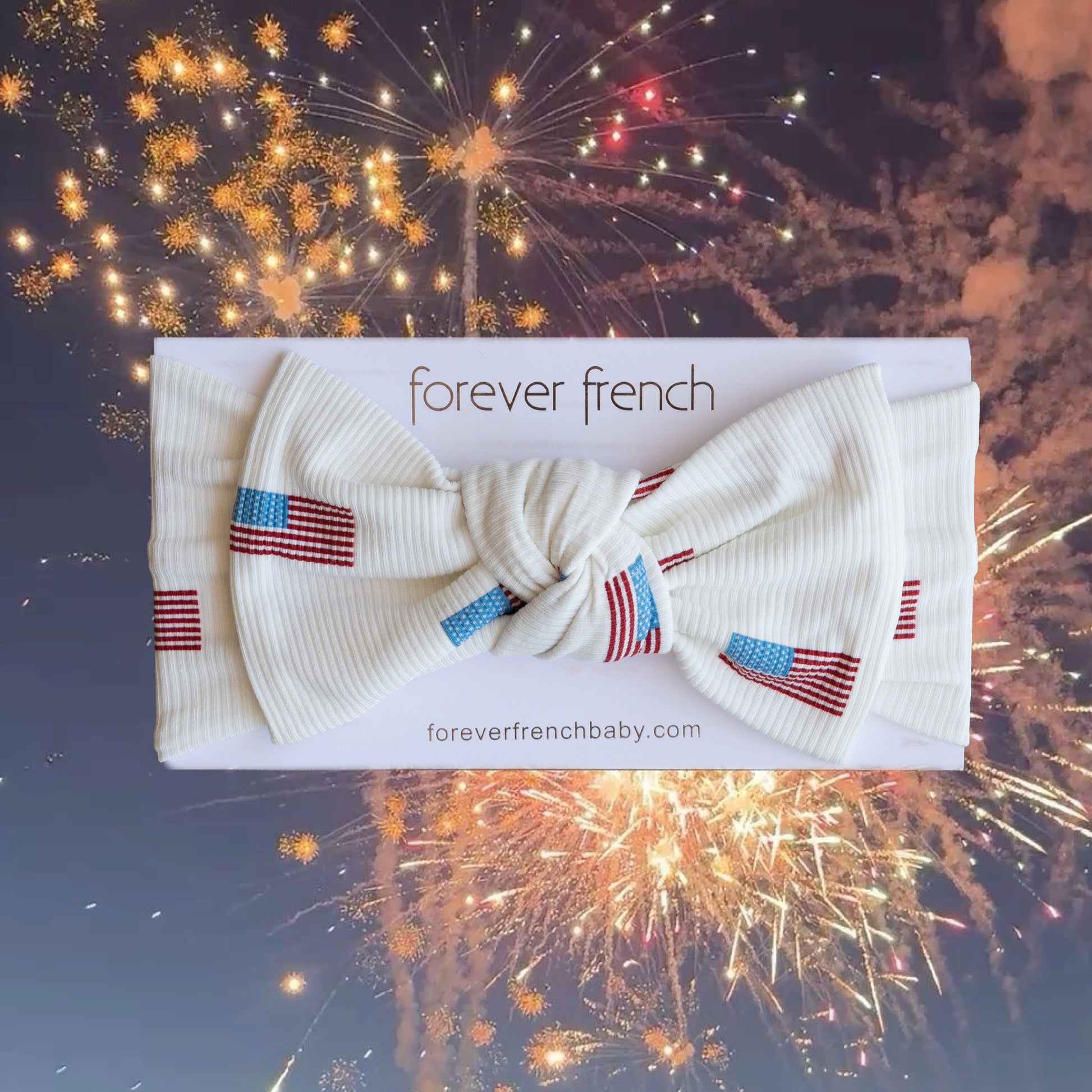 The Bamboo Head Wrap | Americana Ribbed by forever french baby, a white adjustable headband with small American flag designs, is displayed on branded packaging against a backdrop of colorful nighttime fireworks.