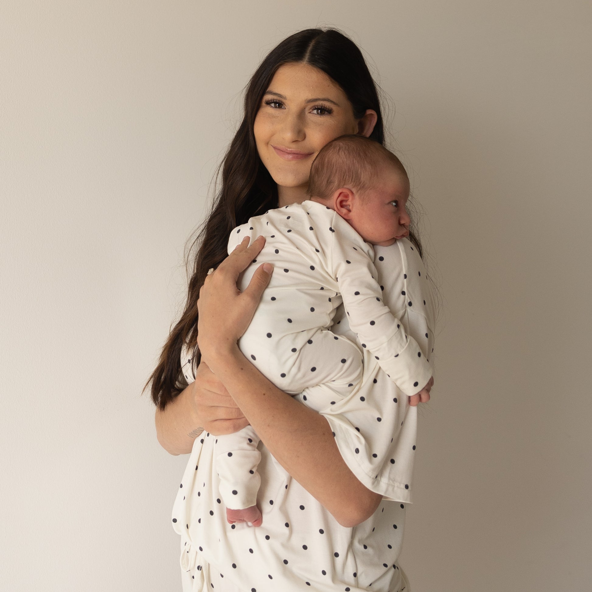A woman with long dark hair smiles while holding a newborn, both wearing the hypo-allergenic Women's Bamboo Short Set | Annie by forever french baby, in front of a plain light wall.